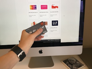 A person holding a credit card in their hand in front of an Apple Mac computer with gift cards displayed on the screen.
