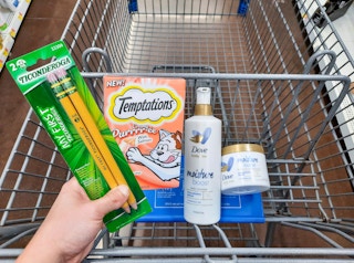 Hand holding Temptations and Ticonderoga products over Walmart shopping cart. Shopping cart has two Dove Body Love products inside it.