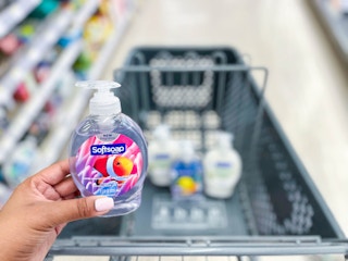 hand holding a bottle of Softsoap hand soap in front of shopping cart with bottles inside it