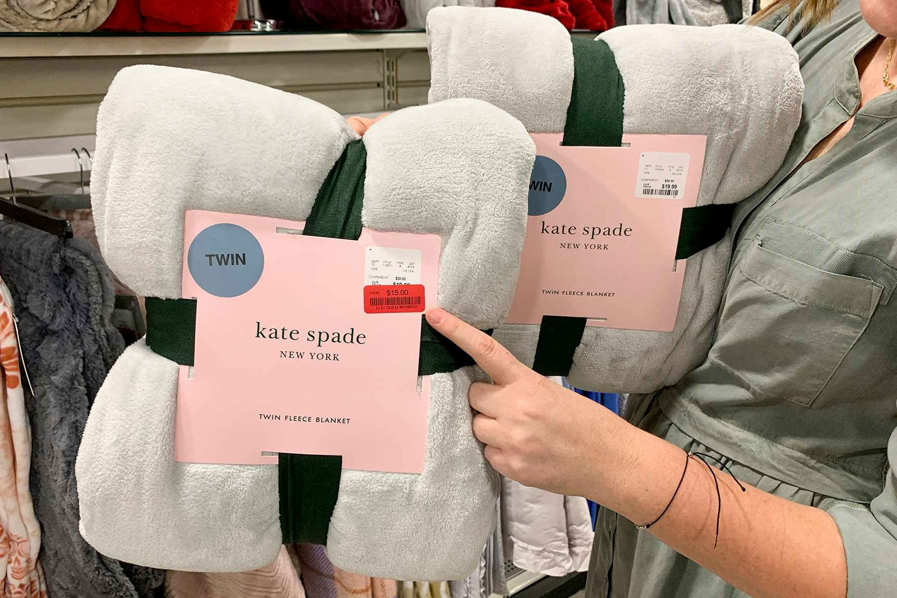 A hand pointing out the sales tag on two identical Kate Spade blankets. One tag shows a red clearance sticker and the other is missing th...