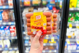 hand holding two packages of oscar mayer wieners in front of store coolers