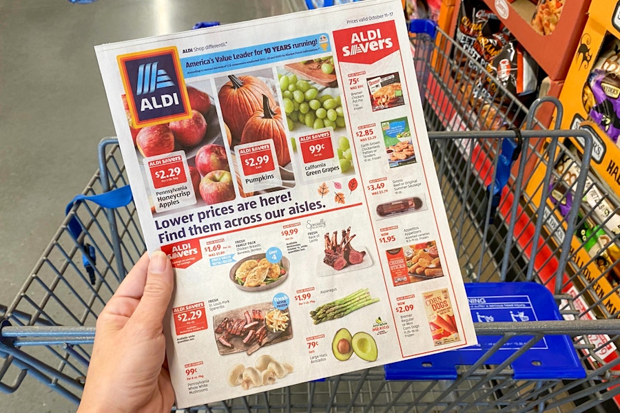 A person holding an ALDI advertisement over a shopping cart inside an ALDI store.