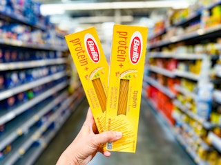 hand holding two yellow boxes of Barilla Protein+ Spaghetti noodles in a Walmart aisle