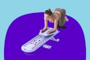 A woman in brown workout clothing leans forward with her elbows on the pilates board and her back arched