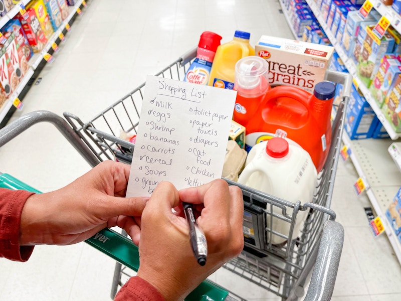 A woman at the grocery store, using a pen to check off an item on her shopping list while resting her hands and wrists on her shopping ca...