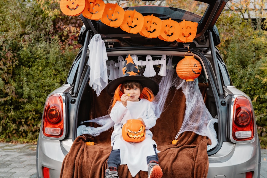 A child dressed in a halloween costume, eating a piece of candy is sitting on in the open truck of a car. The car trunk is decorated with...