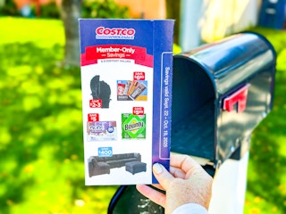 A costco savings book being held up in front of a mailbox 