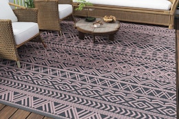 B0DHZDP681 Pink and grey outdoor rug on a deck with some woven rattan furniture with white cushions