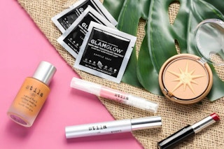 Makeup, lip gloss and other beauty products scattered on a pink and burlap surface.