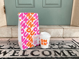 A Dunkin takeout bag and small Dunkin Donuts coffee sitting on a welcome mat outside of someone's front door.