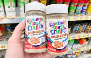 two bottles of cinnamon toast crunch cinnadust held in a hand in front of seasonings at walmart
