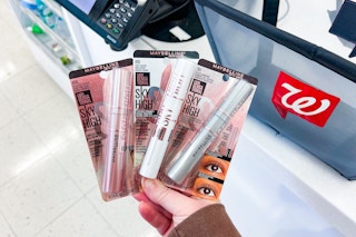 hand holding three maybelline sky high mascaras next to walgreens shopping basket
