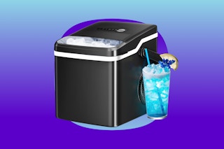 A black countertop ice maker with a thin window in the top and an iced blue drink next to it.