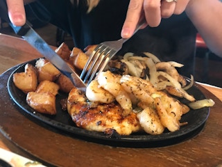 A person's hands using a fork and knife to cut into some chicken on a skillet plate of chicken, shrimp, potatoes, and onions, on a table ...