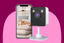 An indoor security camera with a square white housing and stand next to a smartphone