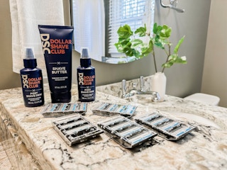 The Dollar Shave Club Shave Butter and two bottles of DSC Post Shave Dew lined up on the counter behind a handful of DSC razor cartridges