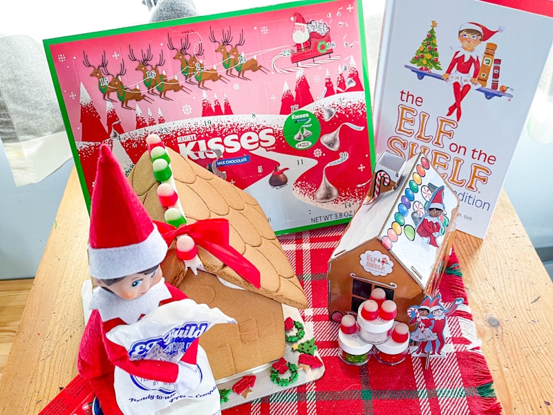 elf on the shelf doll holding frosting next to a gingerbread house, a little cardboard house, book, and advent calendar 