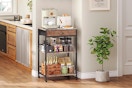 A brown rolling coffee bar cart sits in a beige kitchen. It has two shelves and a drawer.