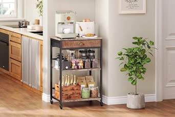 A brown rolling coffee bar cart sits in a beige kitchen. It has two shelves and a drawer.