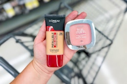 person holding maybelline foundation and blush over a cart