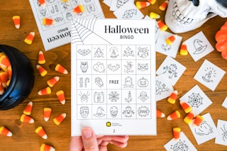 A person holding up a free printable halloween bingo card with candy corn and halloween decor in the background.