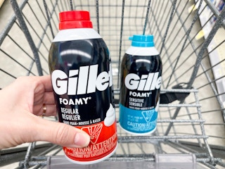 A bottle of Foamy Gillette shaving cream held out by hand in front of another Gillette shaving cream sitting in a shopping cart.