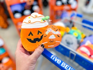 a person holding a squishmellow pumpkin in front of a five below cart 