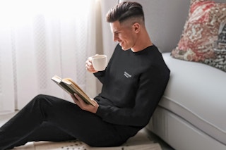 Man sitting up against a couch drinking a cup of coffee wearing the Xtreme Heat Compression Pants & Shirt in black