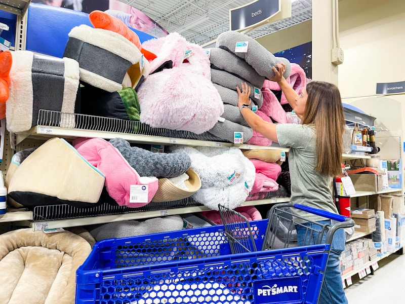 A woman standing next to a PetSmart shopping cart, pulling a cat bed from a stack of them sitting on a shelf at PetSmart.