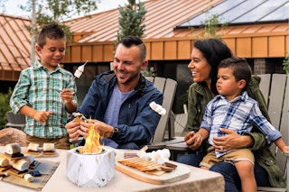 A family of 4 sits at a table outdoors and roasts marshmallows over the tabletop fire pit.