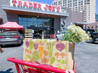 A Trader Joe's shopping cart with bags of groceries in the basket, parked outside of a Trader Joe's grocery store.