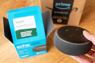 A hand holding an echo dot next to the box it came in and an amazon box.