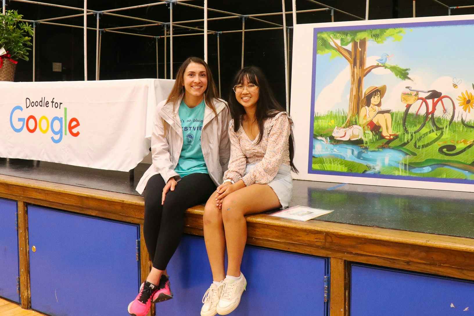 Two students sitting on a stage in front of a student's winning art piece for the state finals of the Doodle for Google scholarship ...