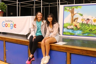 Two students sitting on a stage in front of a student's winning art piece for the state finals of the Doodle for Google scholarship ...