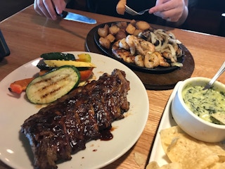 A plate of ribs and vegetables on a table next to a skillet plate of shrimp, chicken, potatoes, and onions, and another plate with a bowl...