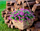 a hanging flower pot with purple artificial flowers in front of wooden logs