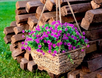 a hanging flower pot with purple artificial flowers in front of wooden logs