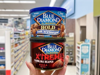 Two Blue Diamond Almond cans held out by hand, stacked on top of each other, in front of a store aisle.