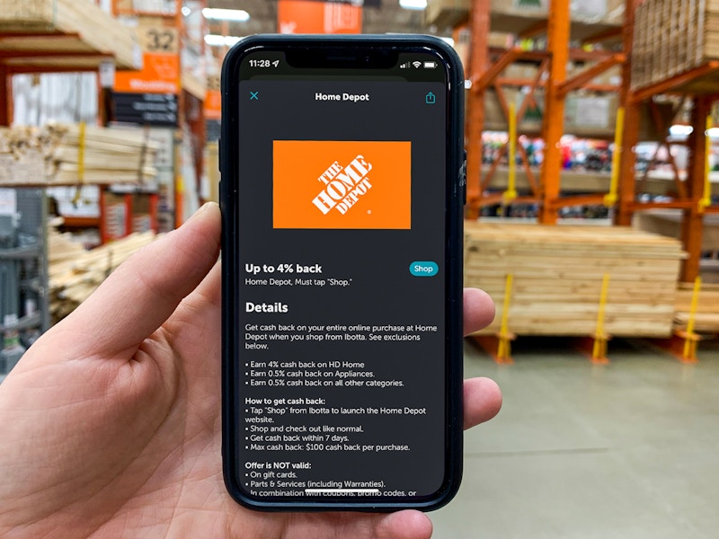 A person's hand holding an iPhone displaying the Home Depot offer page page on the Ibotta app in an aisle at Home Depot.