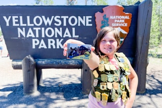 A child in front of the yellow stone national park sign wearing a vest covered in junior ranger badges. The child is holding out a free ...