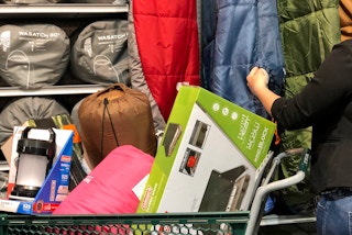 A person looking through sleeping bags next to a shopping cart filled with camping gear including an air mattress and sleeping bags.