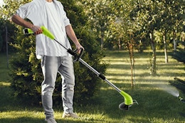 A weed wacker used by a person on a grassy area with several types of trees