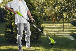 A weed wacker used by a person on a grassy area with several types of trees