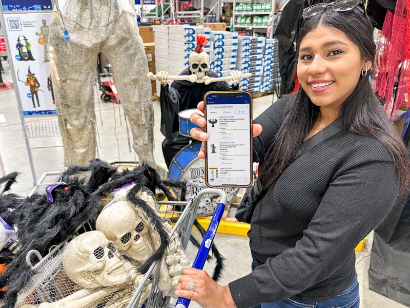 a woman holding out a cell phone with lowes halloween on screeen while holding a cart with decor in basket 