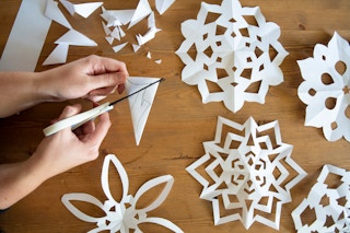 A person cutting a paper to make a snowflake with other paper snowflakes on the table.