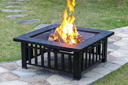 A 32-inch square fire pit has a wood fire burning in it as it sits on a stone walkway in a grassy yard