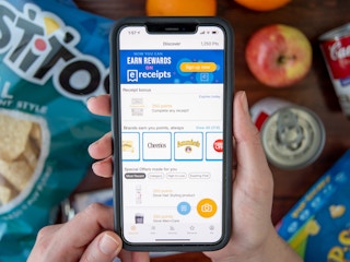 A person's hands holding a cell phone displaying the Fetch Rewards app, with groceries laid out on a table below it.