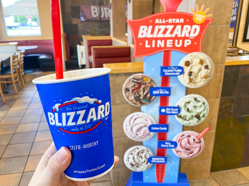 A person's hand holding up a Blizzard cup next to the All-Star Blizzard Lineup cardboard cutout displayed inside Dairy Queen.