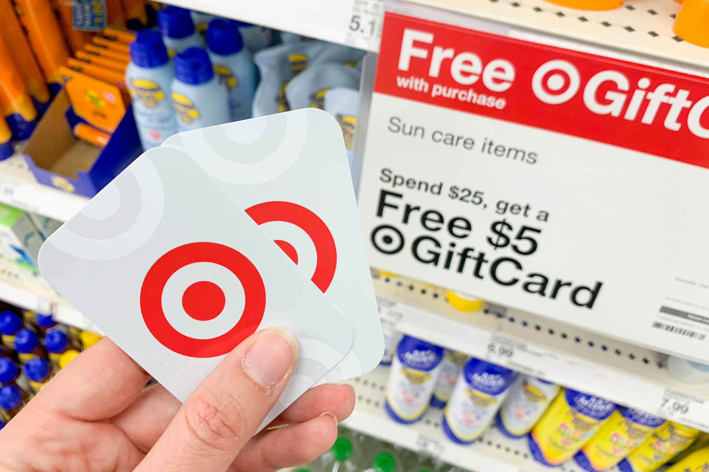 A person's hand holding two Target gift cards in front of a sign advertising getting a free Target gift card when you spend $25 on sun ca...