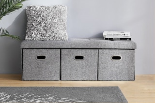 A gray bench with three storage drawers in a room with a fluffy pillow on top